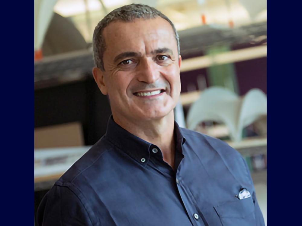 Portrait of a person with short dark hair wearing a navy button-down shirt, standing indoors in front of a blurred industrial or architectural background with curved white structures.