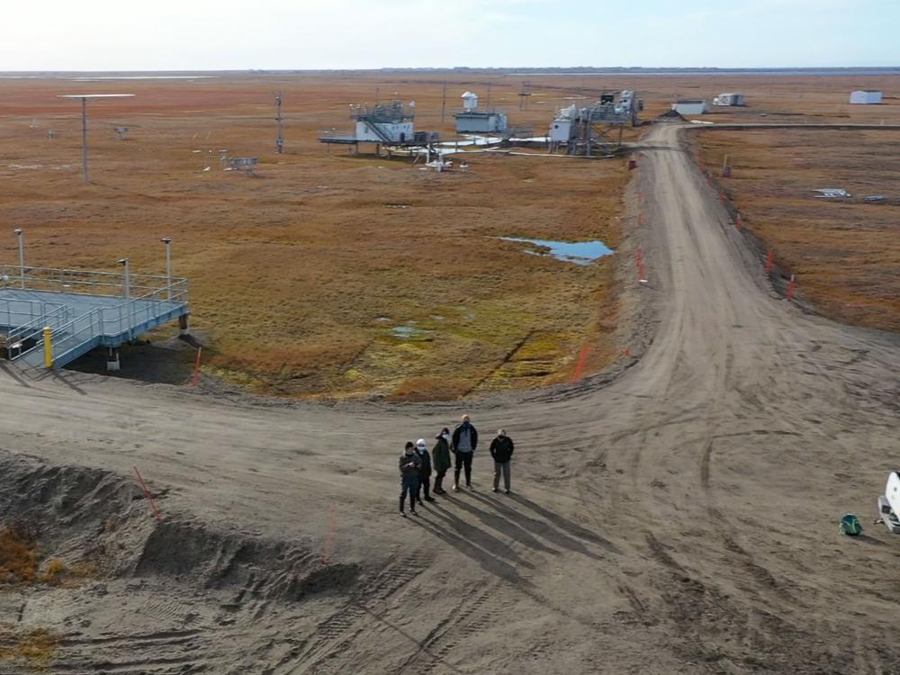 A drone photo showcasing a group of five researchers standing on a road embankment in front of a research facillity in the vast, flat envioronment of Northern Alaska.
