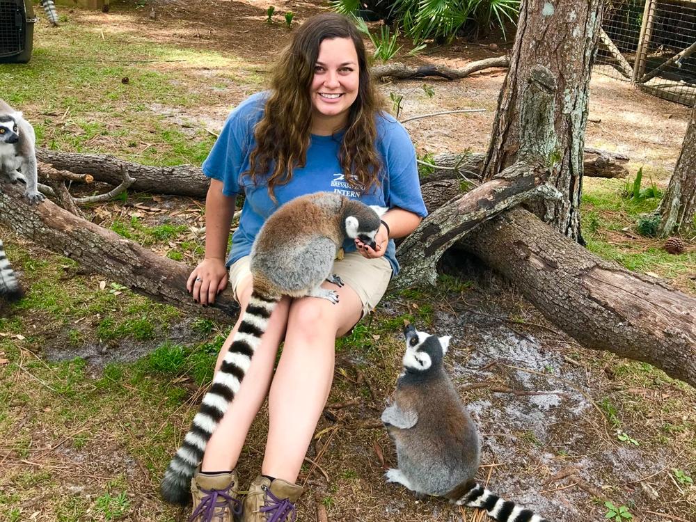 A photo of Nicole Day sitting on the ground with a lemur in her lap and other lemur sitting next to her