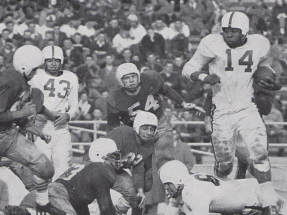 Several football players in dated uniforms are captured in mid-play while a crowd sits in the bleachers in the background.