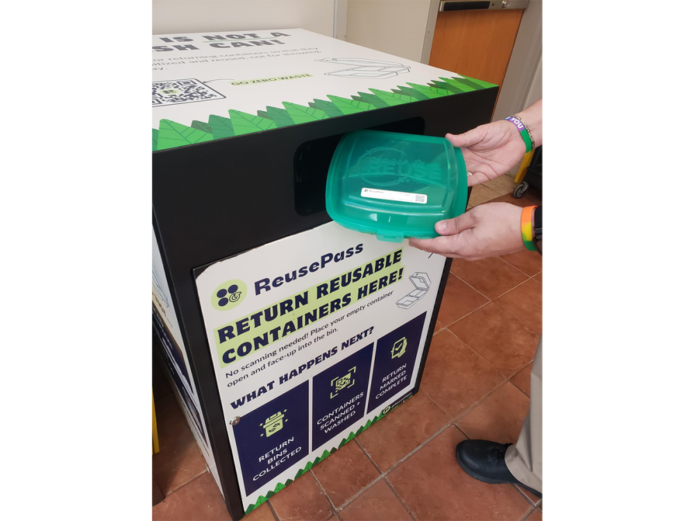 A person returns a reusable takeout container to a collection bin
