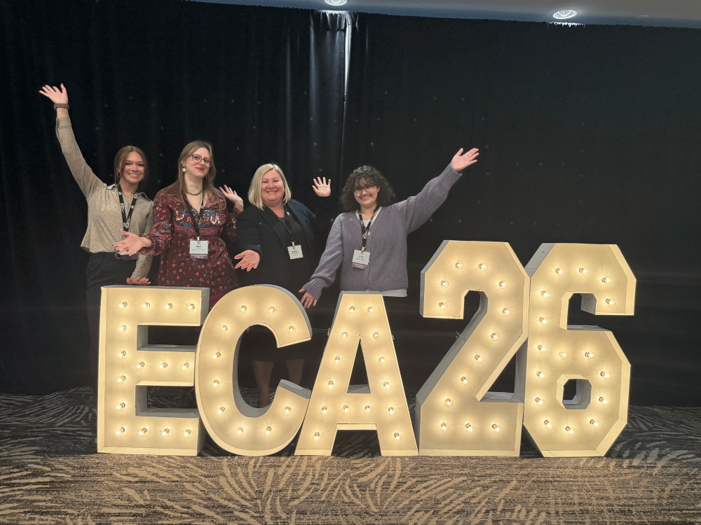 Students and faculty member with their arms outstretched near a large floor sign, "ECA 2026," the conference the group attended