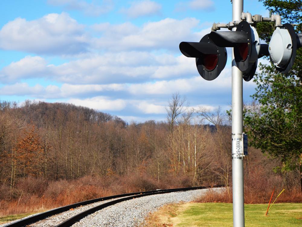 Railroad crossing signal stands beside curving tracks under a blue sky with clouds and leafless trees.