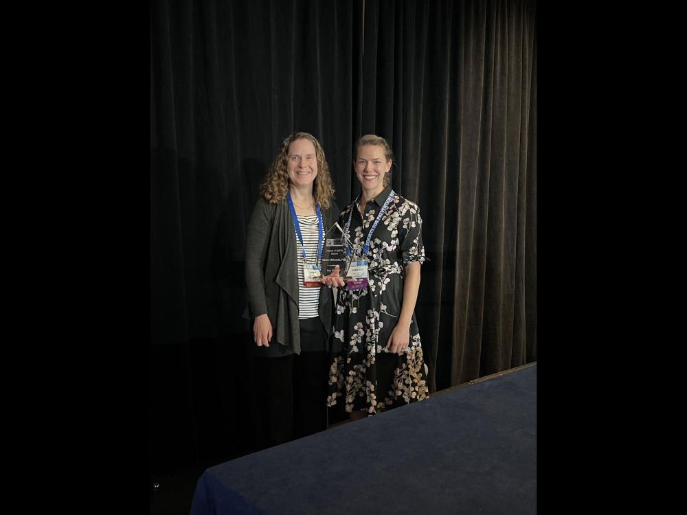 Two smiling women in professional attire, with one holding an award statue