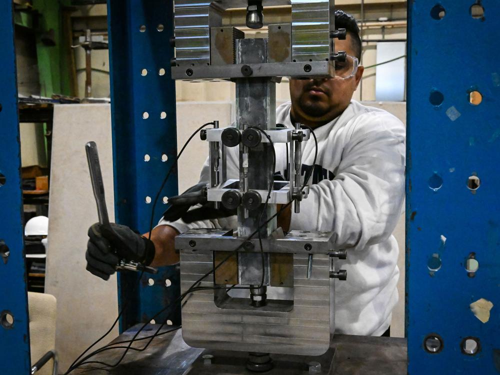 A researcher in a white lab coat and safety glasses operates a large mechanical testing machine in a laboratory to test the strength of concrete