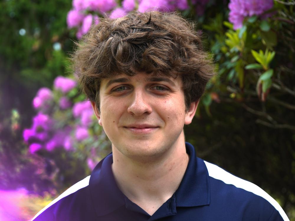 Male student wearing blue and white collared shirt smiles. Purple flowers fill the background of photo.