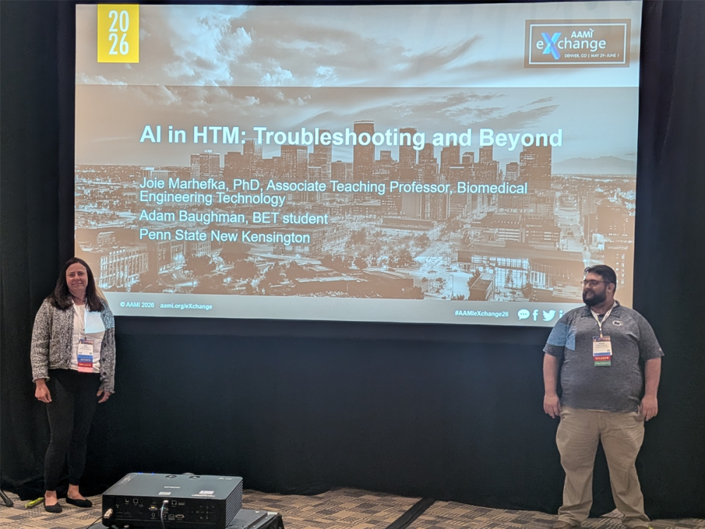 Joie Marhefka and Adam Baughman stand on opposite sides of a projected presentation slide titled “AI in HTM: Troubleshooting and Beyond” at the 2026 AAMI eXchange.