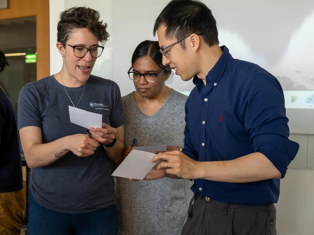 Three faculty members stand in a classroom and compare notes on index cards.