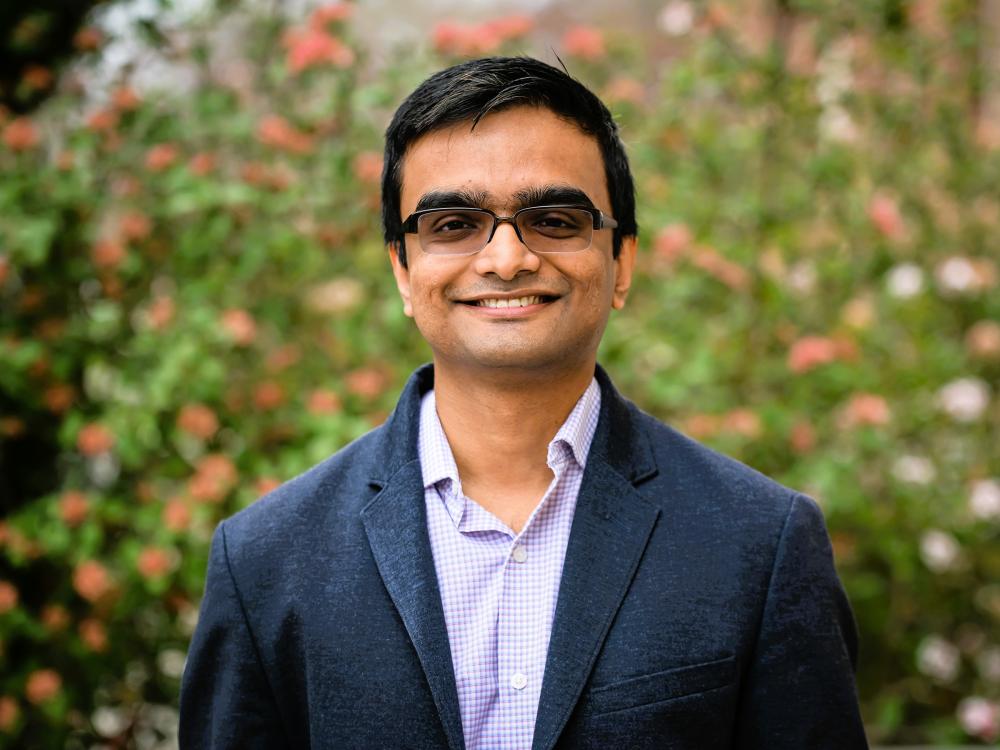 Abhronil Sengupta smiles in a portrait standing in front of a flower bush.