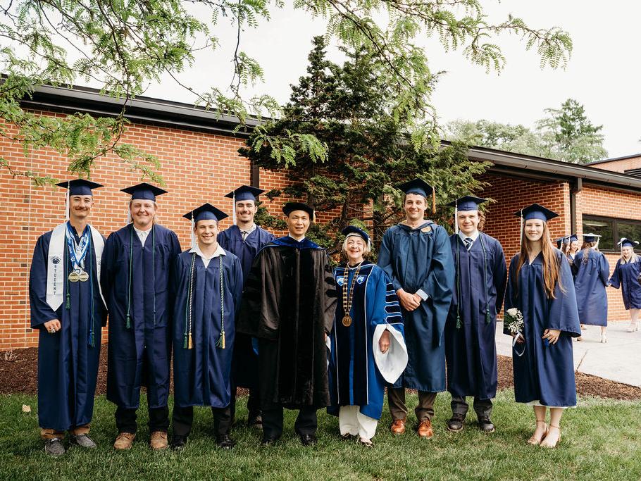 Six graduating members of Penn State Mont Alto's championship Woodsmen Team stand outdoors with Linghao Kong, professor of chemistry and interim director of academic affairs, and Marilyn J. Wells, regional chancellor of Penn State Brandywine, Penn State Mont Alto and Penn State York. All are dressed in commencement regalia and pose in front of a campus building.