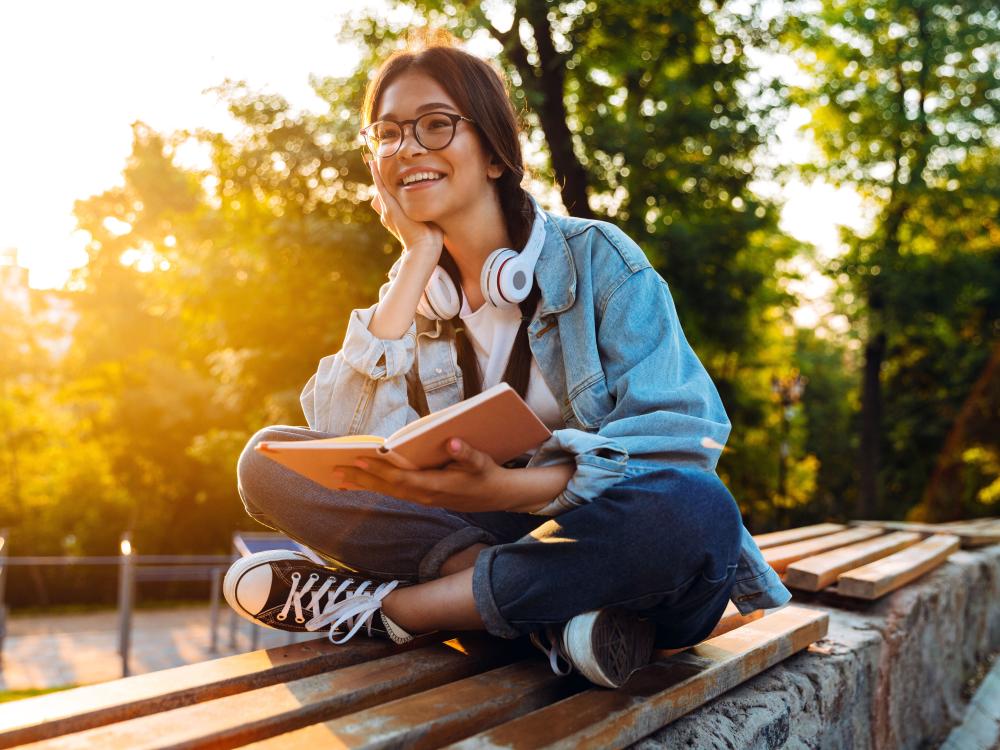 A smiling teenager sits outdoors on a bench reading a book on a sunny summer afternoon.