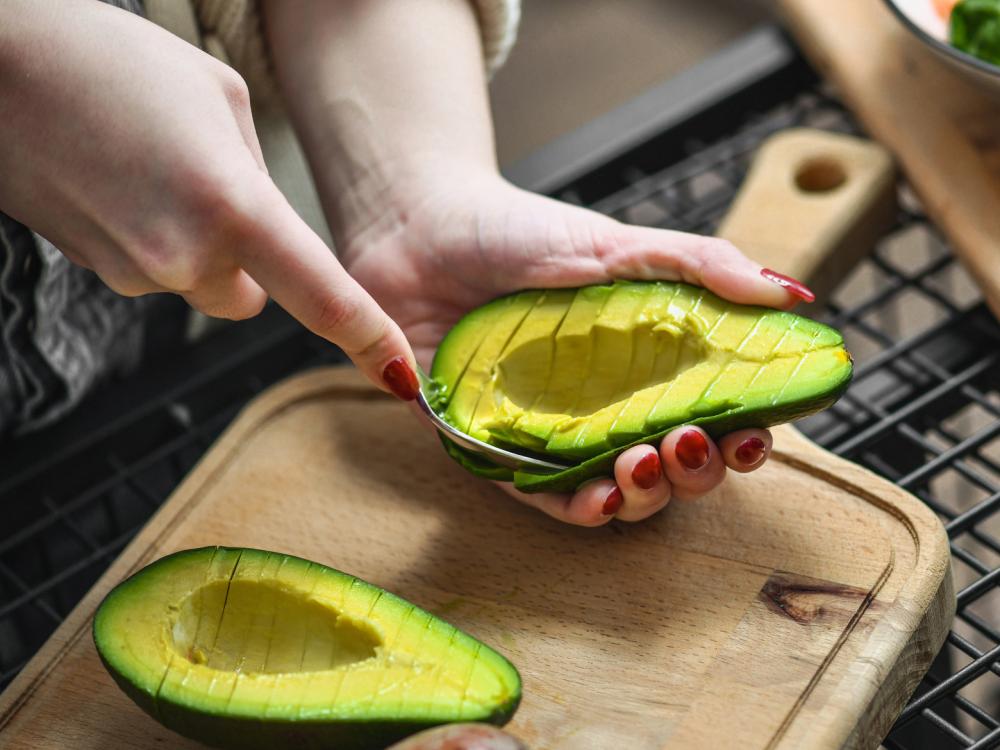A woman's hands scoop a ripe avocado above a cutting board in a kitchen