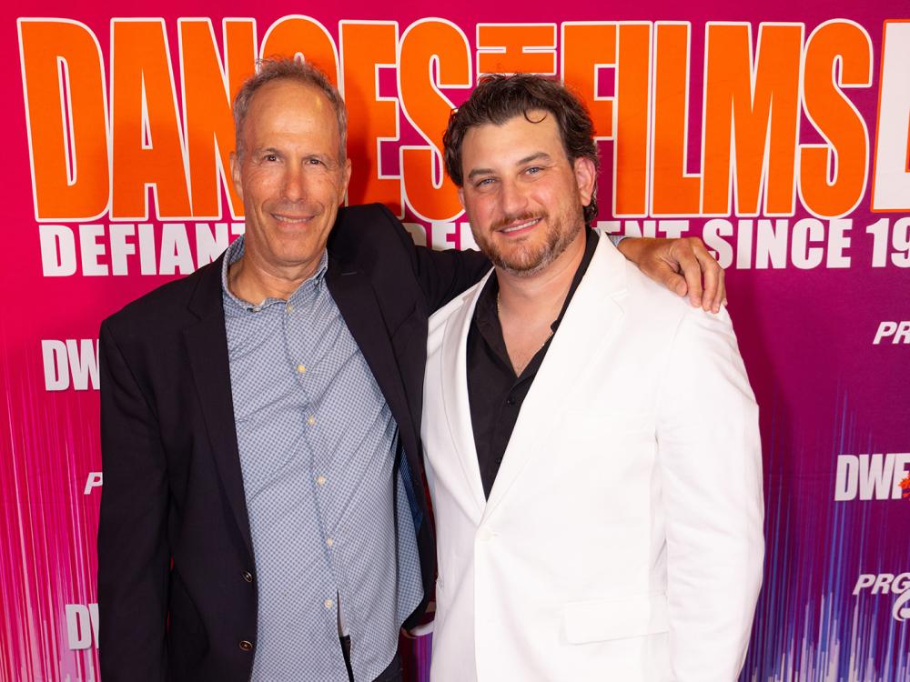 A man in a dark coat and shirt with his arm around a dark-haired man in a white coat.