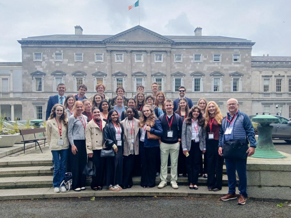 Group of adults posing in front of a historic government building with an Irish flag overhead on a cloudy day.
