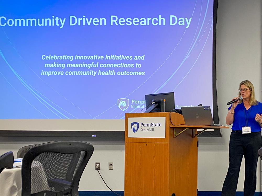 Miriam Kelly Miller speaks into a microphone at a wooden podium. Behind her, a large projection screen displays the text "Community Driven Research Day" alongside the Penn State Clinical and Translational Science Institute logo.