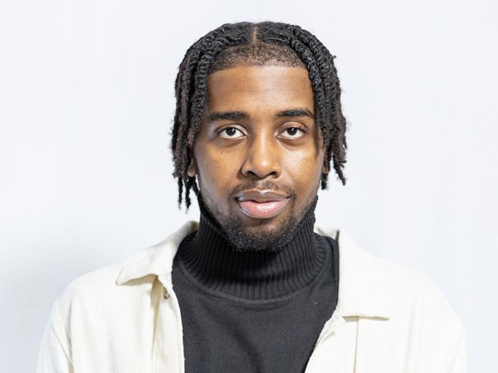 A black man with dark curly hair, wearing a light-colored coat.