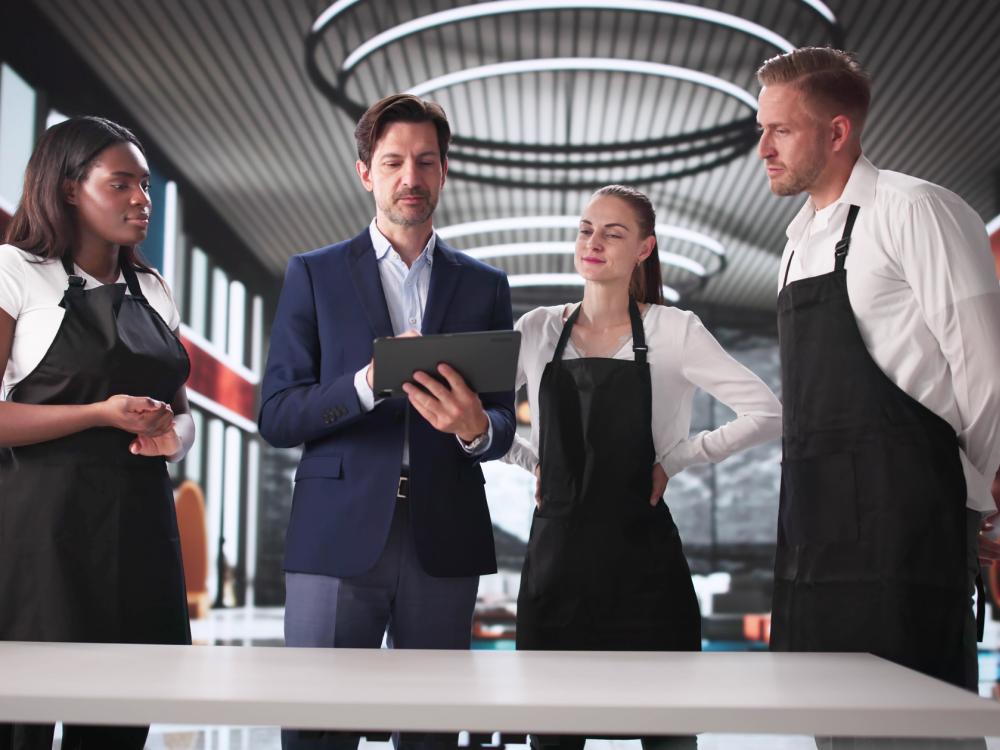 A manager and three wait staff stand in an otherwise empty restaurant, discussing tasks for the coming shift.