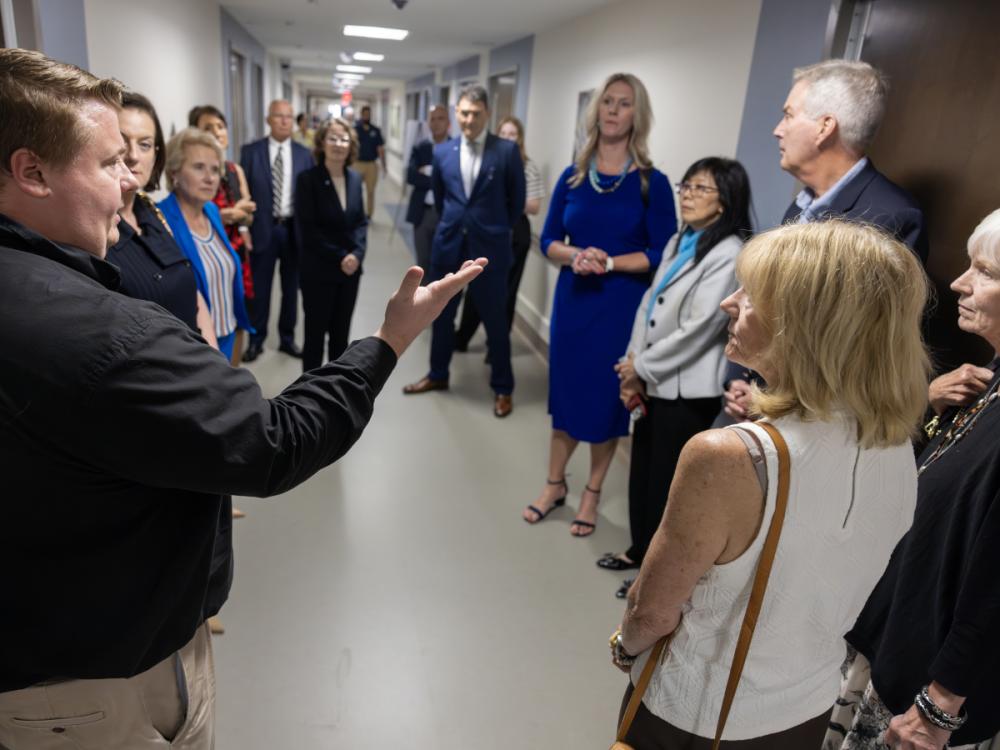 A group of about 15 people wearing professional attire stand in the hallway of a newly refurbished hospital unit. A man in the foreground has his arm extended outward as he speaks to the group.