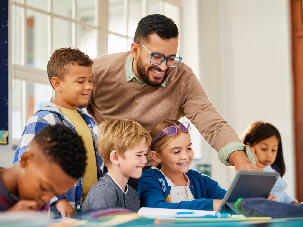 An elementary school teacher smiles as he leans over a table to help a small group of students using a tablet during class, while other children work independently in the bright, modern classroom.
