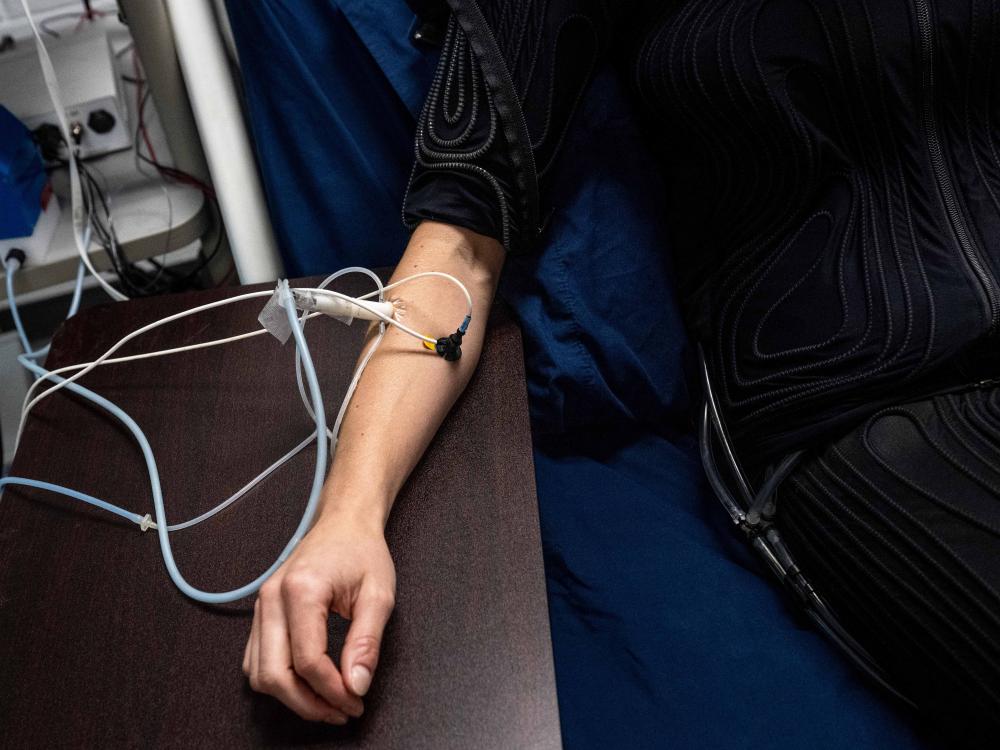 The torso of a woman in a black suit covered with small hoses reclines on a hospital bed. Her right arm rests on a table with two sensors attached to it.