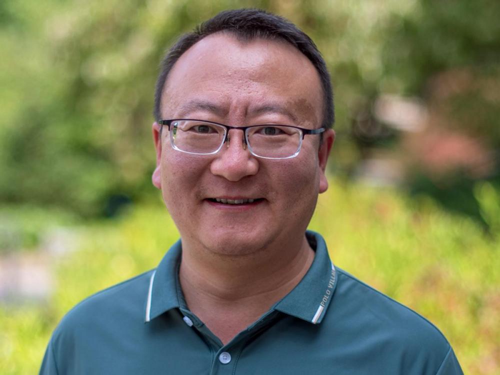 Zhenlong Li, wearing glasses and a green collared shirt, smiles outdoors with blurred greenery in the background.