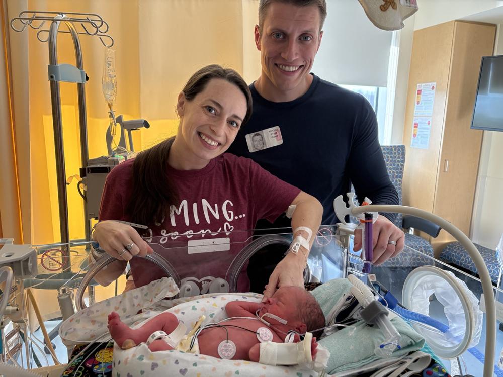 A mother and father smile with their infant son who is in an incubator in a hospital neonatal intensive care unit. The mother touches the baby’s hand.