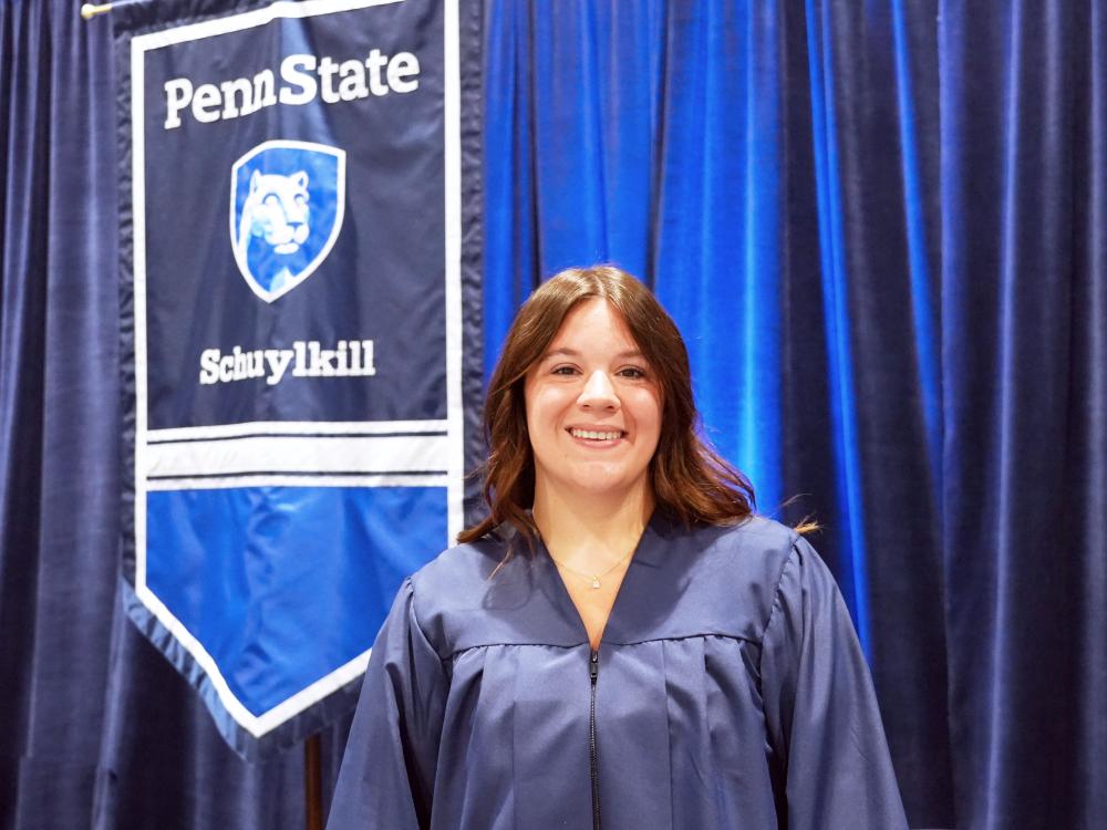 Student stands in front of flag with the Penn State Schuylkill logo in her graduation gown