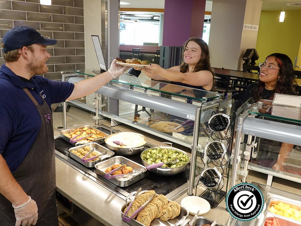A staff member at Pure Dining Station in the East Food District serves food to two female students (AllerCheck Seal included)