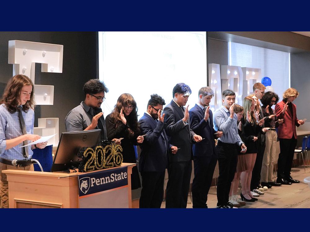 student inductees stand in front of room in single line with hands raised to take the Engineering Oath