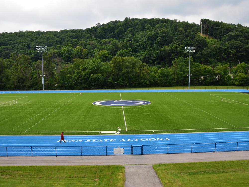 Penn State Altoona athletic field with a blue track, green turf, and wooded hillside in the background.