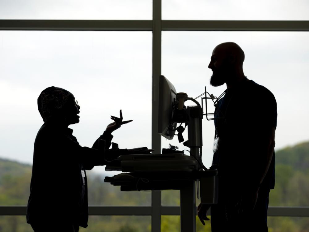 A silhouette of two employees standing and talking in front of a window at Penn State Health St. Joseph Medical Center. A computer on a cart is between the two of them.