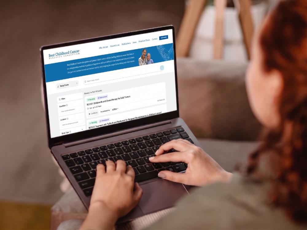 A view from over the shoulder of a person typing on a laptop computer. On the screen is a page within the Beat Childhood Cancer Research Consortium website that displays Study Finder search results.