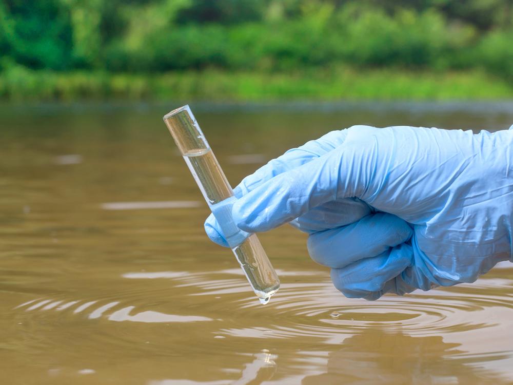 a gloved hand holds a beaker of water above a body of water