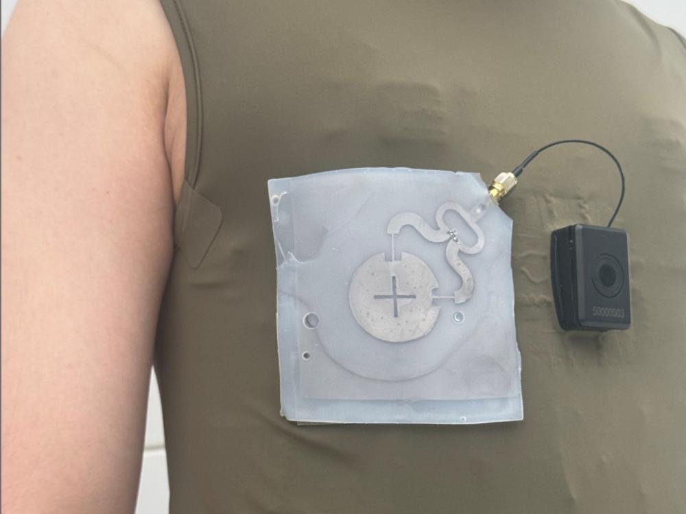 Close up of a person's chest, wearing a olive green sleeveless shirt, with a sensor attached to it.