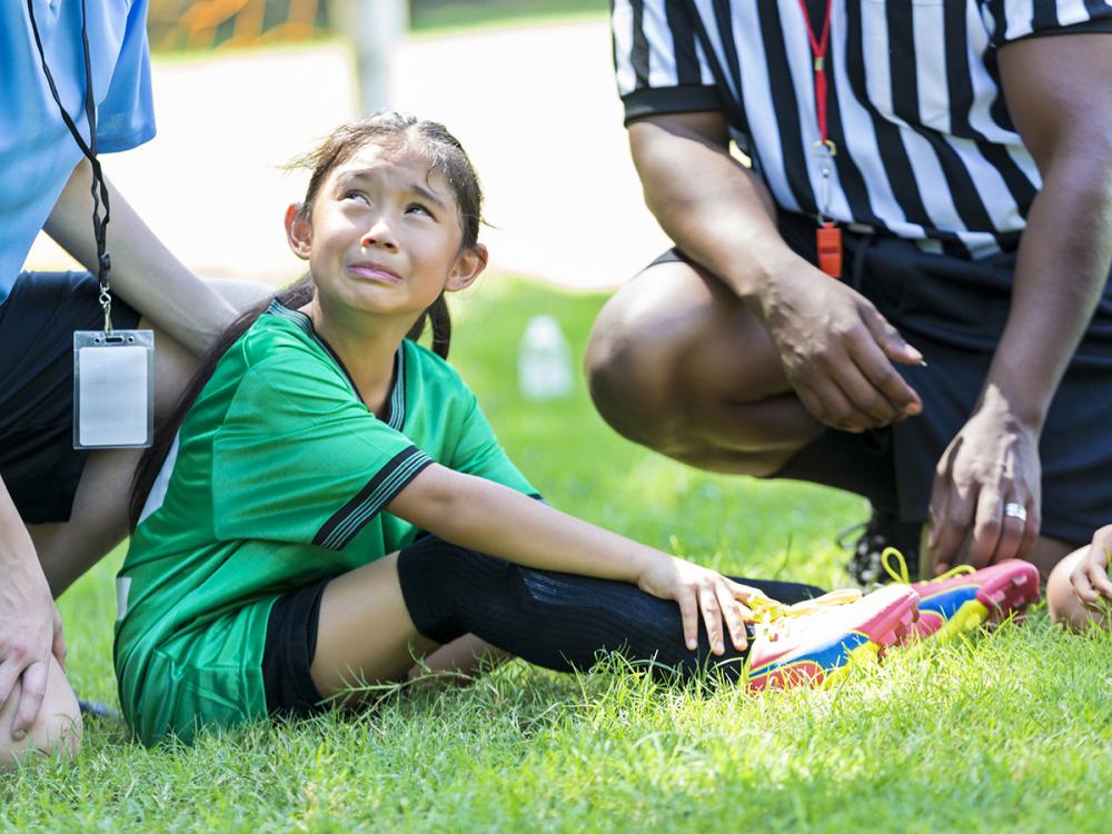Young female soccer player holds her ankle and grimaces in pain while crying. She is sitting on the playing field with her coach and referee gathered around her. 