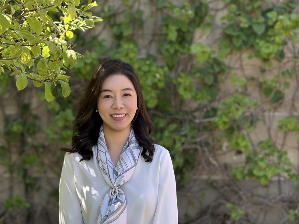 A woman with shoulder-length brown hair smiles and stands in front of green vegetation.