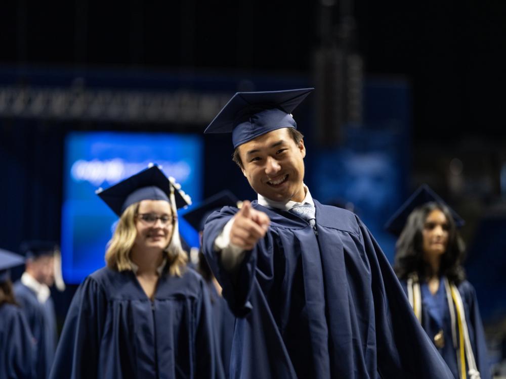 A person wearing a blue cap and gown smiles and points at the camera