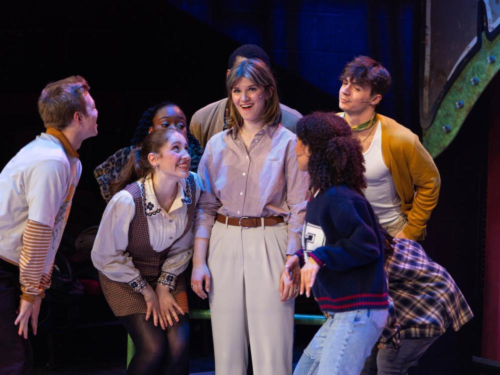 A woman in blouse and slacks surrounded by actors/singers