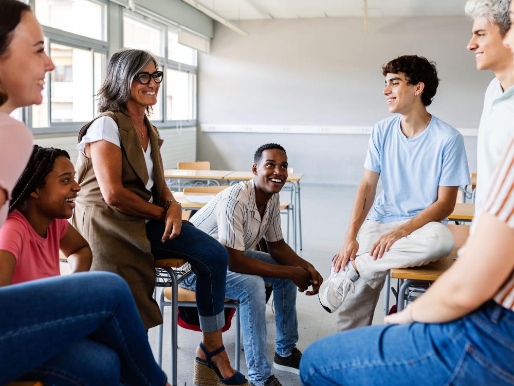 A diverse group of high school students and educators sit together in a classroom, smiling and engaging in an informal group conversation.