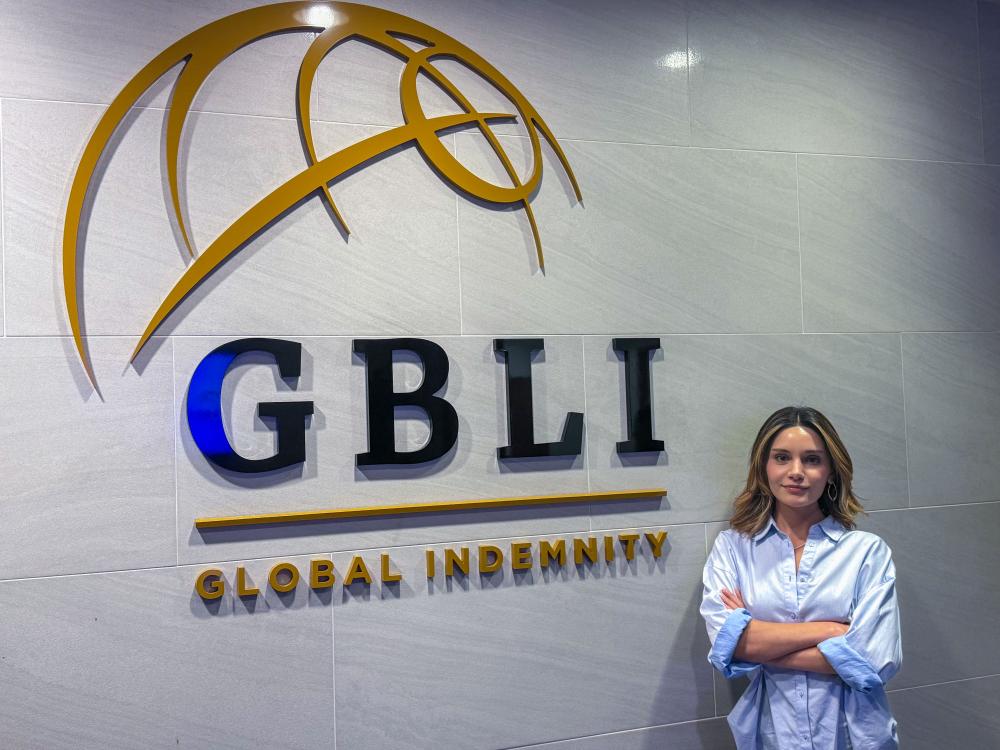 Emine Barik posing in a light blue shirt stands with her arms crossed in front of a corporate logo that says "GBLI" in black and blue and "GLOBAL INDEMNITY" in gold.