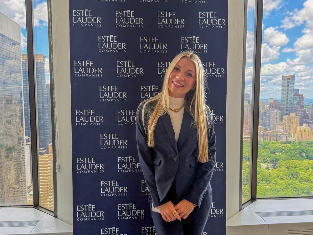 Karli Brenneman wears a black suite while standing in front of a banner that reads “Estée Lauder Companies” with the New York City skyline out the window next to her.