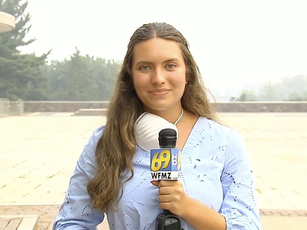 A young woman with long brown hair holding a microphone stares at the camera, 