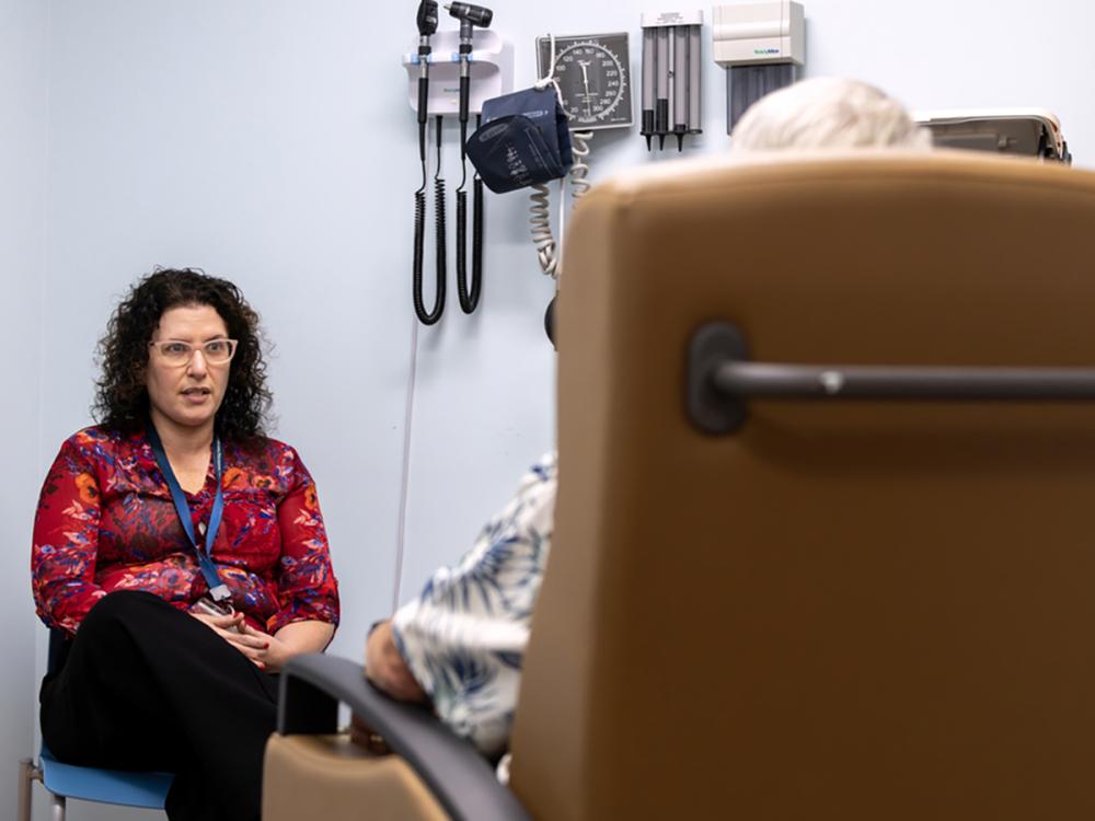 Sarah Kawasaki talks to a patient seated in an office exam chair. The patient is shown from behind. A blood pressure cuff and other medical instruments hang on the wall nearby.