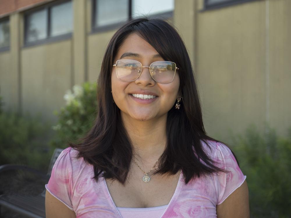 Female student with glasses smiles in front of brick building.