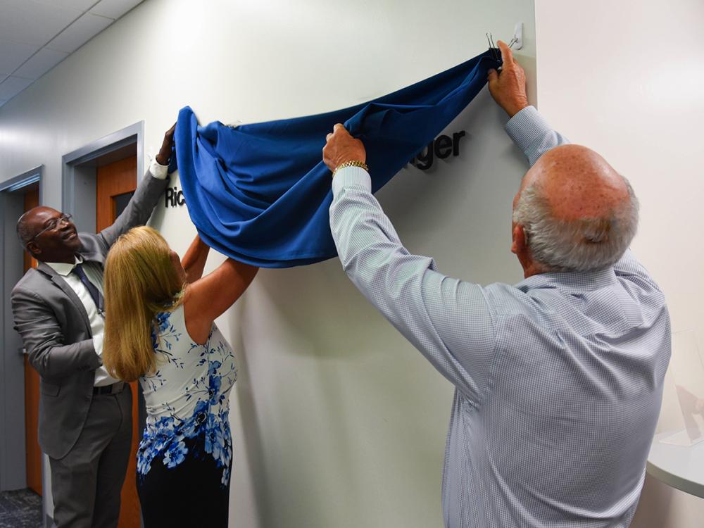 Ron Darbeau, Jeane Singer, and Richard Singer unveil a plaque in a hallway.