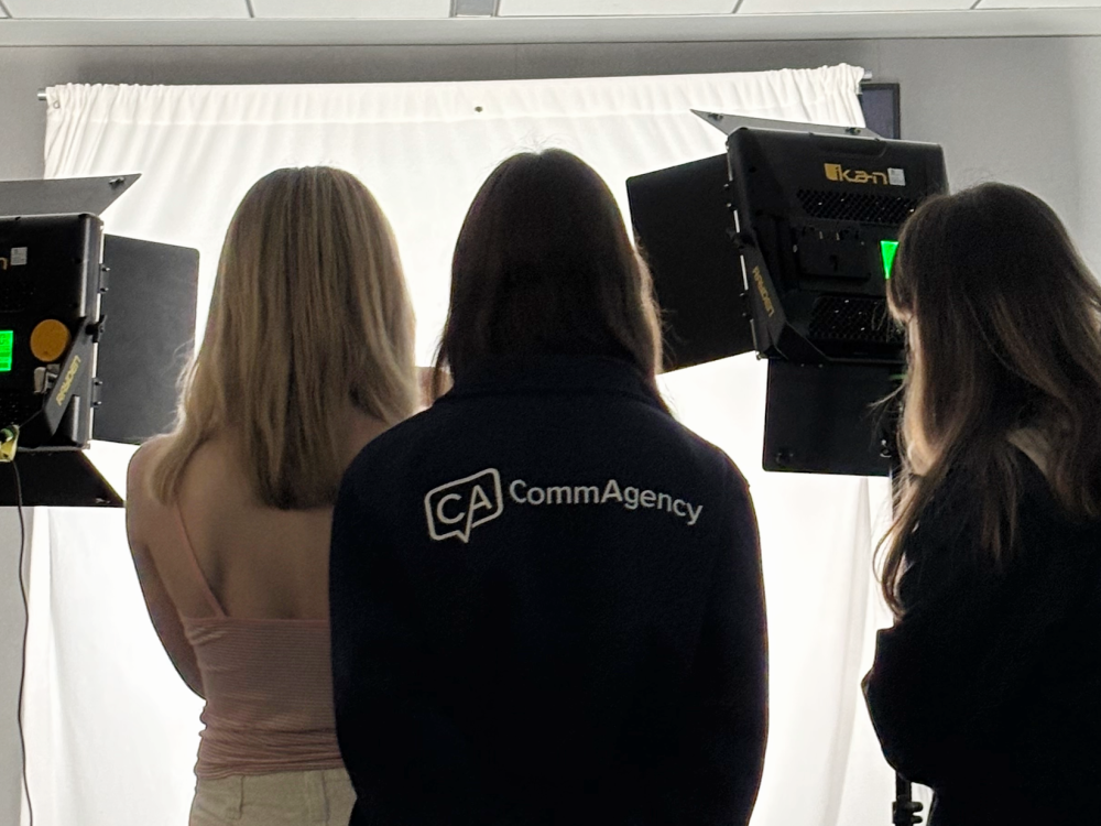 Three students stand with their backs to the camera, with light stands beside them for a photo shoot. One student is wearing a shirt that says "CommAgency" on the back.
