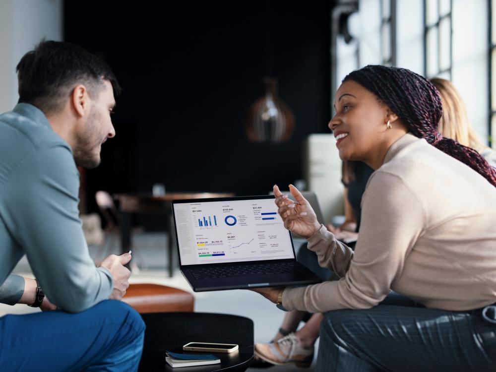 Two people sit in a well-lit, public, indoor space, looking at charts on a laptop screen.
