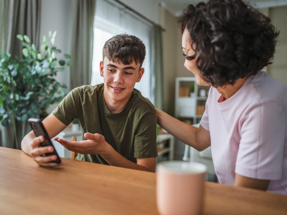 A pleased teenage boy discussing something on his telephone with his smiling mother, who rests her hand on his shoulder.