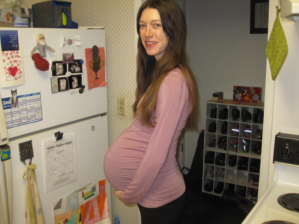 Heidemarie Laurent smiles at the camera while holding her pregnant belly and standing in a kitchen.
