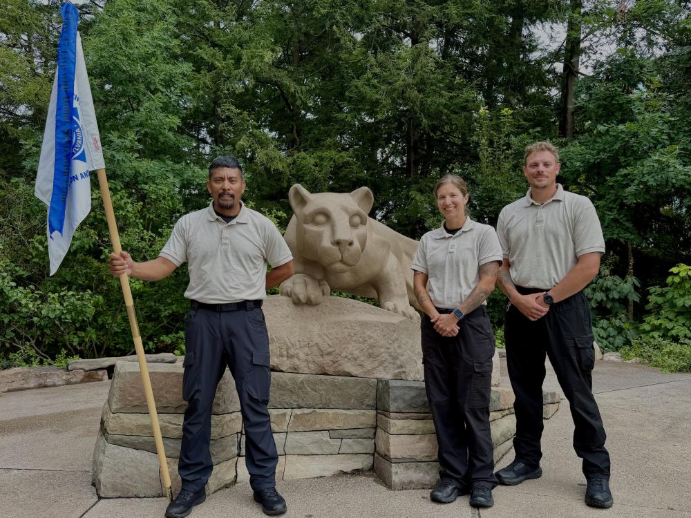 three people with a blue and white flag at the nittany lion statue